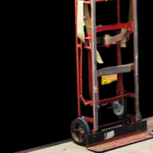 A red dolly with visible wear, as though it's been used as rental equipment, is positioned on a concrete surface against a black background. The dolly has two wheels and a vertical handle structure with a few straps hanging loosely.