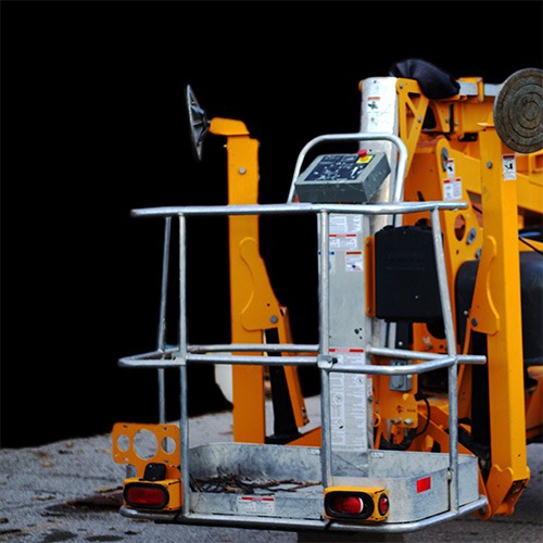 A yellow cherry picker with a metal safety cage, control panel, and stabilizing elements stands on a concrete surface against a dark background. The equipment is used for aerial work.