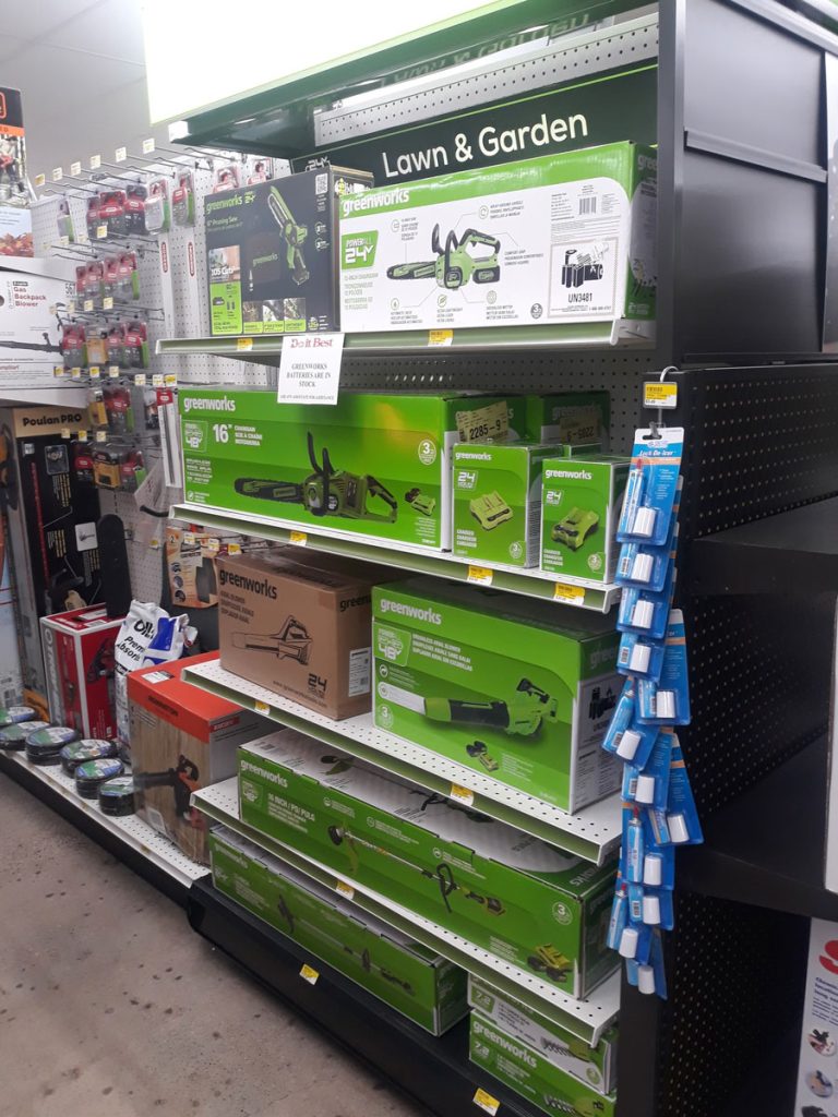 A Carlisle Hardware aisle with a display of lawn and garden tools, featuring green boxes with various equipment like trimmers and chainsaws. Shelves are stocked with products, and there are small hanging items on the right. A "Lawn &amp; Garden" sign is visible.