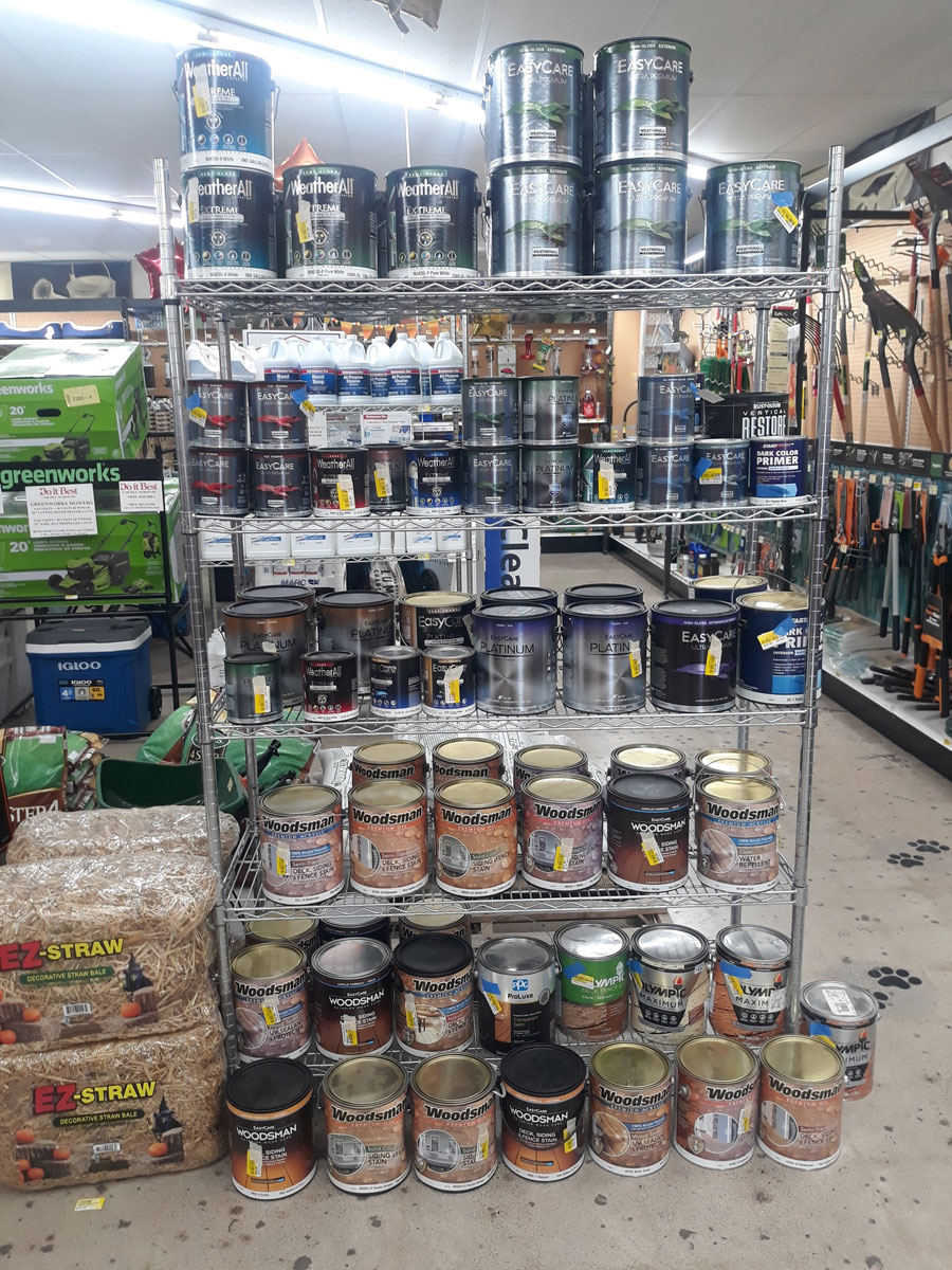 A store shelf at Carlisle Hardware features various cans of paint and wood stain. The top shelves boast gallon cans labeled "Weatherall" and "Skycote," while the bottom holds smaller cans marked "Woodsmart." EZ-Straw bags rest on the floor, with gardening tools visible in the background.