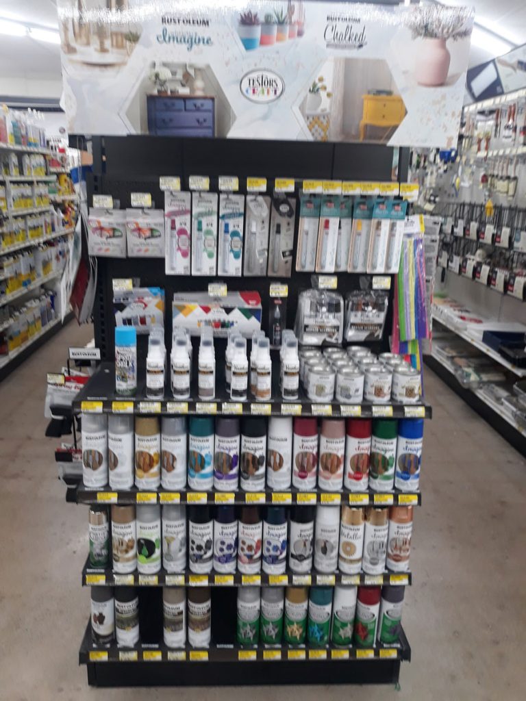 The display rack at Carlisle Hardware holds various spray paint cans and supplies in a bustling store aisle. The top banner showcases different paint projects, while the shelves are stocked with colorful cans and painting tools, with labels and prices clearly visible for an easy shopping experience.