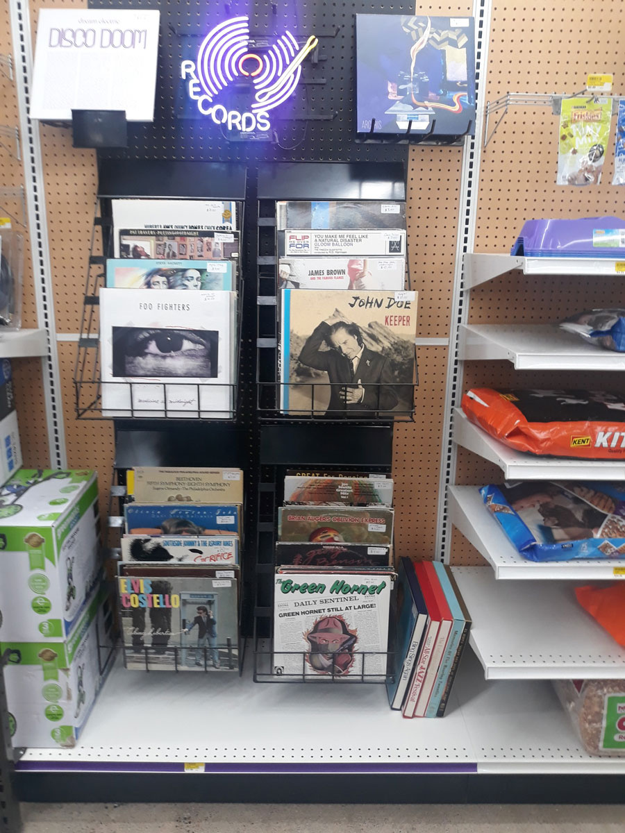 A store display at Carlisle Hardware features vinyl records on metal shelves, showcasing albums from artists like Foo Fighters and John Doe. A glowing purple "Records" sign sits above. Nearby, shelves hold pet food and various other essentials.