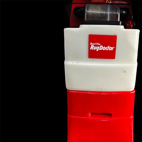 Close-up of a red and white Rug Doctor carpet cleaning machine against a black background. The machine features a prominent red sticker with white text that reads "Rent The Rug Doctor.