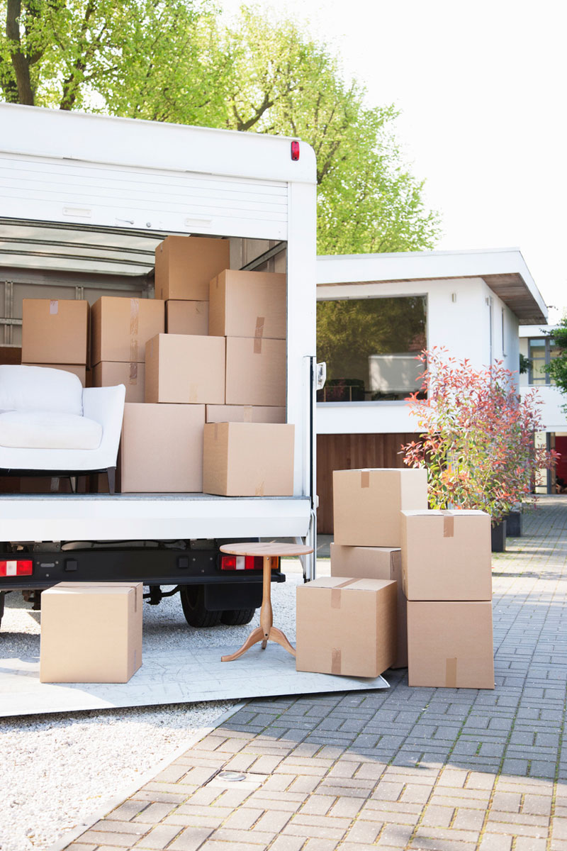 A moving truck is parked on a driveway with its back door open, revealing several stacked cardboard boxes and a white sofa. More boxes are on the pavement nearby. A modern house and garden are in the background, under a clear blue sky.