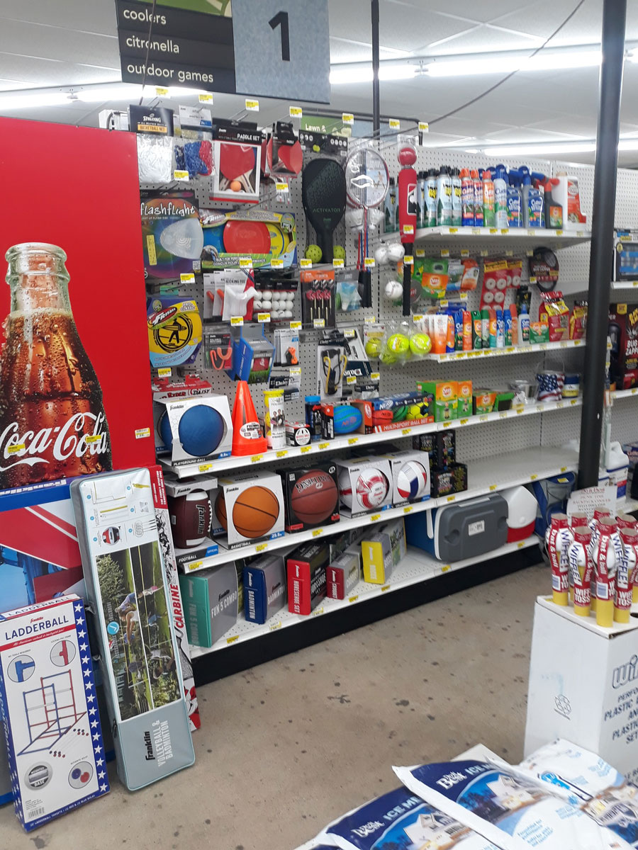 Store aisle at Carlisle Hardware displaying sports equipment and recreation items, including basketballs, footballs, tennis balls, and various games. Shelves are stocked with bottles and cans on the side. A large Coca-Cola display is visible on the left.