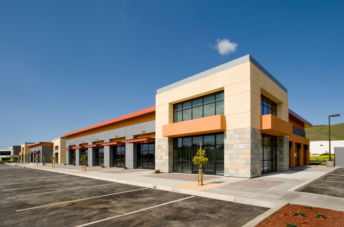 Empty commercial building with large glass windows and stone accents under a clear blue sky. The parking lot is vacant, and small shrubs are planted along the walkway.