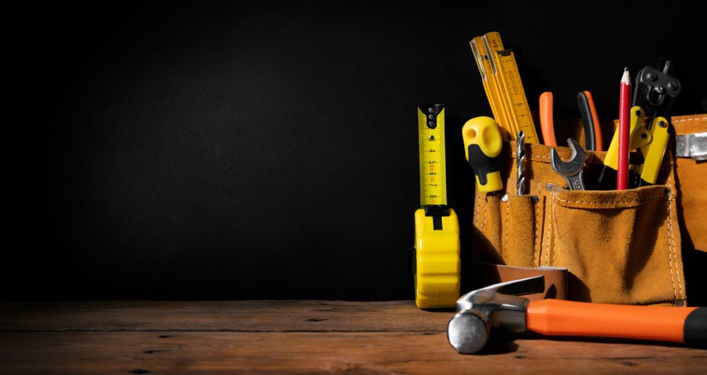 A selection of tools, including a tape measure, pliers, wrenches, and screwdrivers, are placed in a brown tool belt on a wooden table against a black background. An orange-handled hammer lays in the foreground.