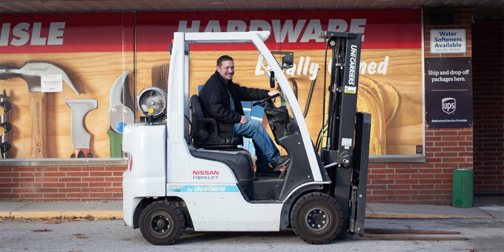 A person smiles while sitting on a white Nissan forklift in front of a hardware store. The store's exterior displays tools and signs advertising various services, including UPS packaging and water softeners.