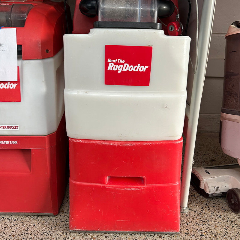 A red and white Rug Doctor carpet cleaning machine is positioned on a speckled floor. The brand logo "Rent The Rug Doctor" is prominently displayed on the front. Another machine is partially visible to the left.