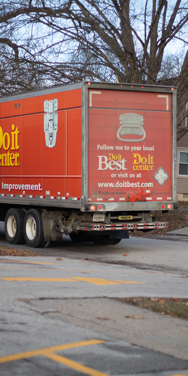 An orange truck with "Do It Best" branding on the back is parked on a street. It has promotional text inviting people to visit local Do It Best centers. Leafless trees and a house are in the background.