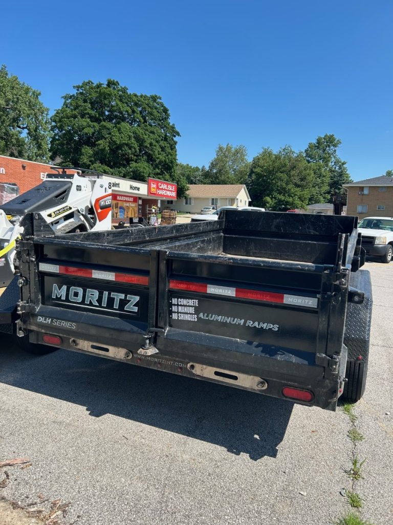 A black Moritz DLM Series dump trailer, ideal for dump trailer rentals, is parked on a paved area near a hardware store under clear blue skies. The trailer’s back gate is open, and equipment can be seen on the left with trees in the background.