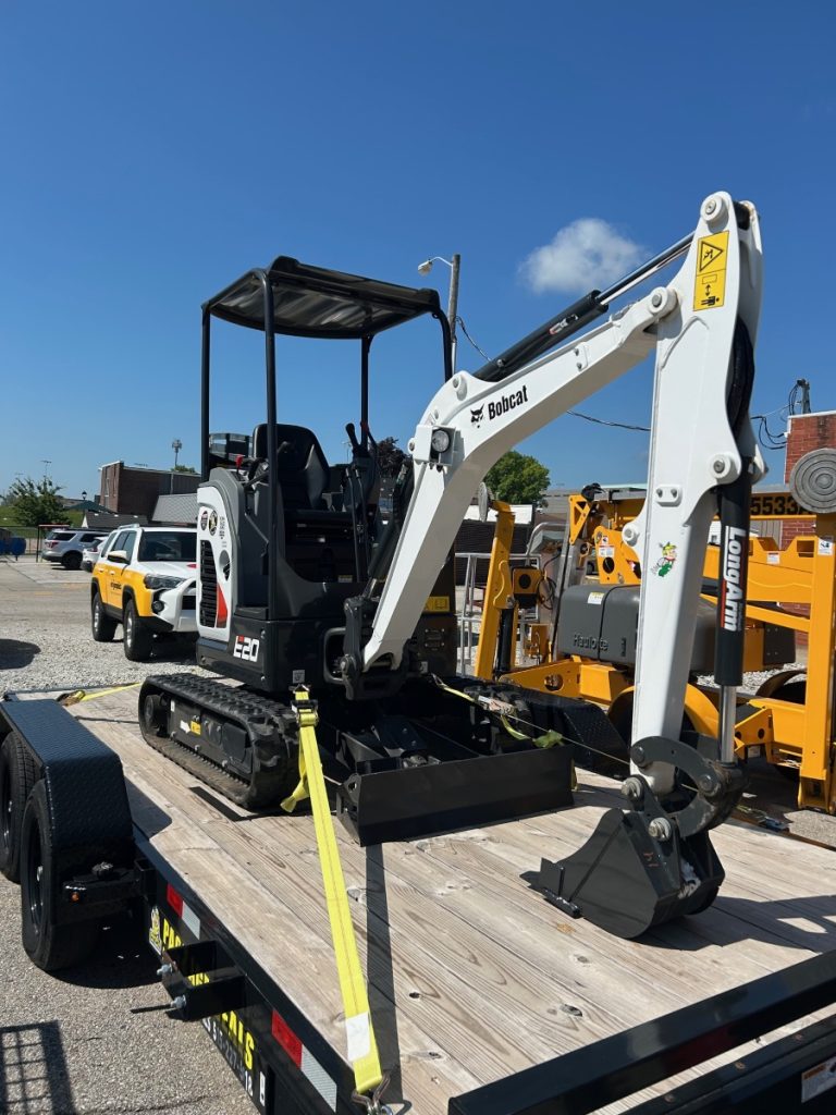 A small white and black Bobcat E20 compact excavator, ideal for excavator rentals, is secured on a flatbed trailer with yellow straps. The background features other construction vehicles and equipment under a clear blue sky.