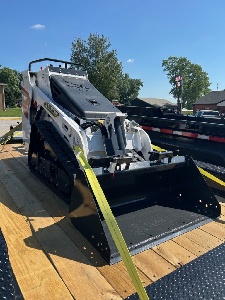 A compact track loader, available through skid loader rentals, is secured with yellow straps on a flatbed trailer. The machine sits on wooden planks under a clear blue sky with some trees and buildings in the background.